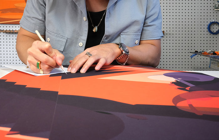 A person in a light blue shirt and jewelry uses a craft knife to meticulously cut paper on an orange and black abstract artwork, reminiscent of Adrian Kay Wongs Cherry Print. The workspace mimics his detailed style, with tools on a pegboard background.