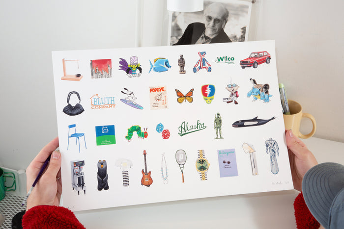 A person holds the Andrew Watch - "Multiples 2" limited edition print, featuring rows of small, colorful, hand-drawn pop culture objects and icons against a white background, above a white desk with a mug, lamp, and a black-and-white photo.