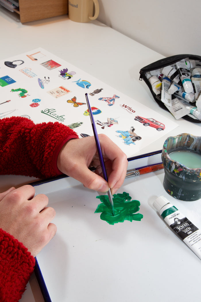 Clad in a red sweater, a person uses a brush to paint a green shape on paper, inspired by the Andrew Watch - "Multiples 2" print from Andrew Watch. Nearby sit a water cup, a tube of green paint, and a sheet filled with vibrant stickers and illustrations.