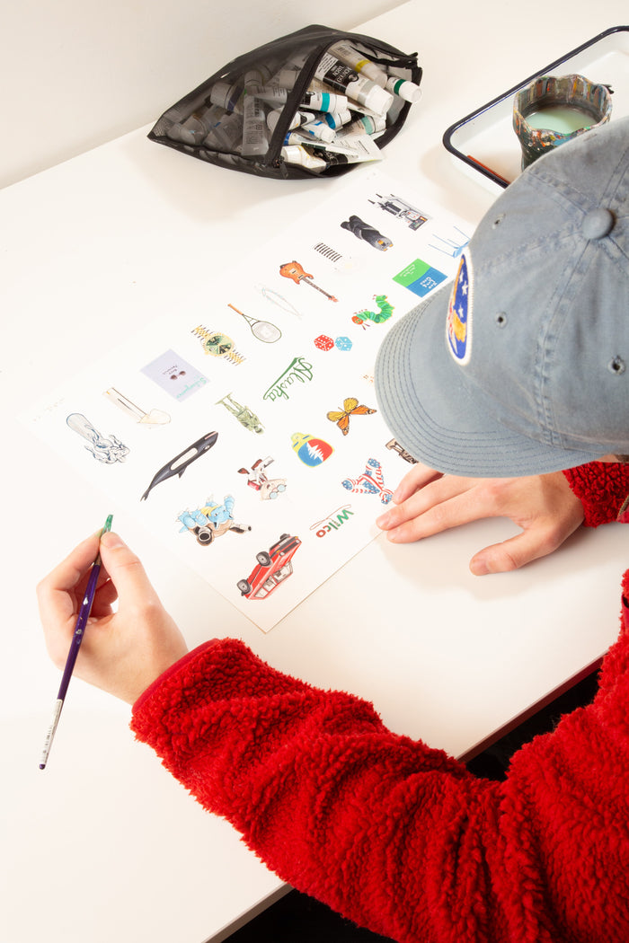 A person in a blue cap and red sweater paints colorful illustrations on paper at a white desk, surrounded by paint tubes, a brush, and a cup—creating vibrant artwork reminiscent of the Andrew Watch - "Multiples 2" print by Andrew Watch.