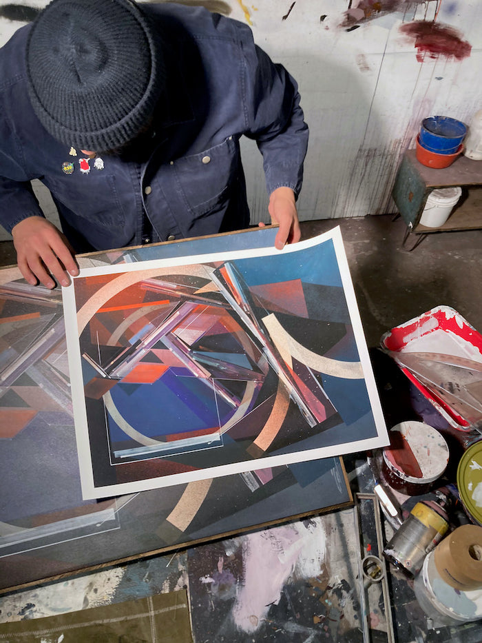 A person in a blue jacket and beanie looks down at "Dodge&Move," an archival pigment print by Augustine Kofie featuring geometric shapes and bold colors, in a studio filled with art supplies. This limited edition piece from Augustine Kofie is signed and numbered, enhancing its exclusivity and artistic value.