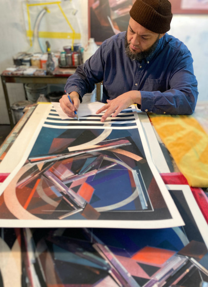 Augustine Kofie sits at a table in his studio, carefully signing and numbering limited edition "Dodge&Move" abstract geometric prints, surrounded by art supplies.