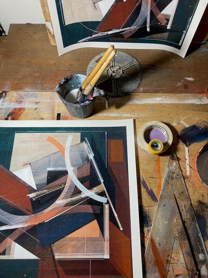 The Augustine Kofie - "Pyledriver" print by Augustine Kofie showcases abstract geometric artwork set atop a wooden table, surrounded by creative tools like a small paint can filled with brushes, rolls of tape, and visible paint splatters. This limited edition print captures the vibrant energy and intricate details of the artist's hand-painted process.