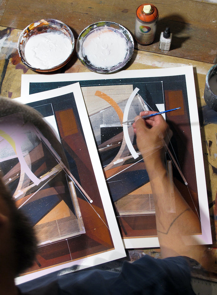 An artist’s hand and arm are visible as they transform two Augustine Kofie - "Pyledriver" prints into unique hand-painted multiples, painting white lines atop each archival pigment print. The abstract geometric prints rest on a table alongside paint supplies, brushes, and a spray can.