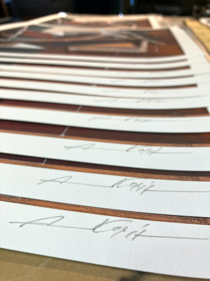 A close-up of several Augustine Kofie - "Pyledriver" prints from Augustine Kofie arranged in a row on a table, each featuring a pencil signature along the white border at the bottom. The focus is on the signatures in the foreground.