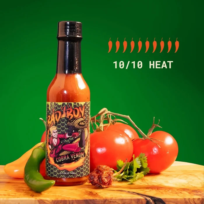 Bad Boy Cobra Venom hot sauce bottle with black cap, surrounded by fresh peppers and tomatoes on wooden surface