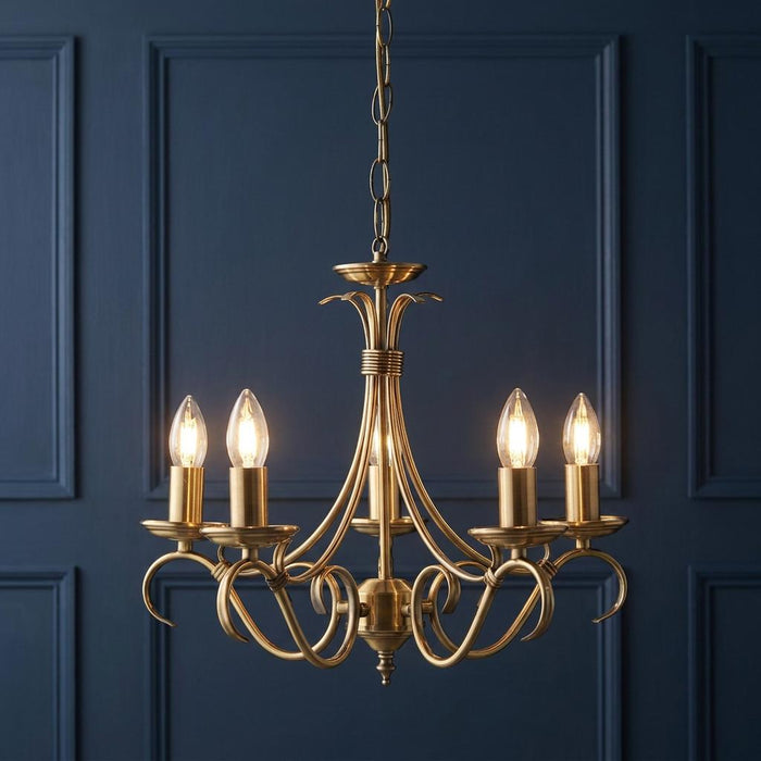 Endon Bernice 5-light antique brass chandelier with lit candle bulbs against a dark blue paneled wall.