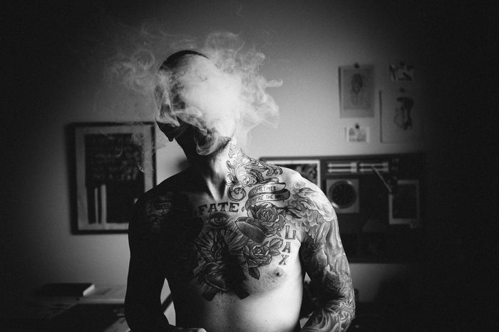 A tattooed individual smokes, their face concealed by swirling smoke, in the moody ambiance of dim walls with framed pictures. This scene is expertly captured in Shaun Roberts limited edition archival pigment print, Black is a Blind Remembering…” (with Ron Ulicny, Portland, OR).