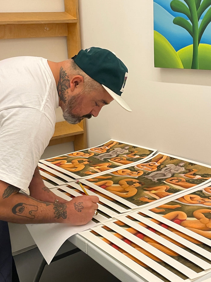 A tattooed individual wearing a white shirt and green cap signs Carlos Rodriguezs limited edition Cupids Garden archival pigment prints in earthy tones on a table, with a framed painting as the backdrop.