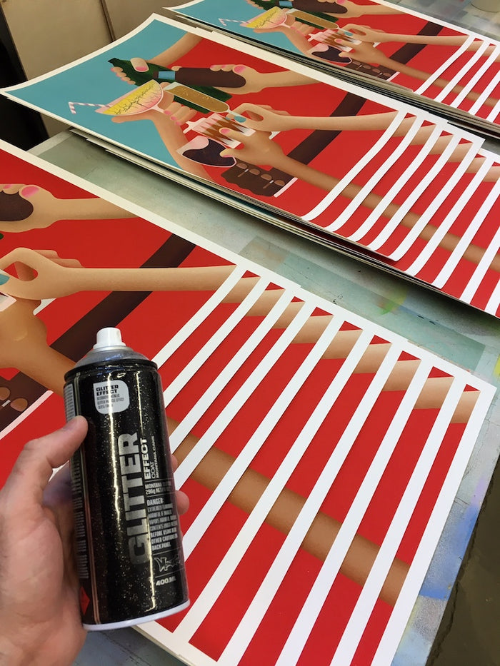 A hand holds a can of black Glitter Effect spray paint above several colorful, limited edition Casey Gray - "Cheers" prints, featuring red and white stripes and illustrated arms holding drinks.
