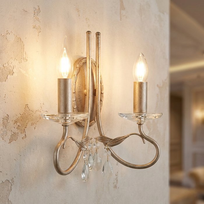 Silver and gold two-light wall sconce with crystal droplets and leaf details lit against a textured wall.