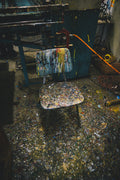 A paint-splattered chair in a cluttered workshop echoes the vibrant chaos of “Chuck’s” by Shaun Roberts with Chuck Sperry. The colorful floor mirrors an archival pigment prints artistry, while tangled cords and industrial machinery complete the lively scene.