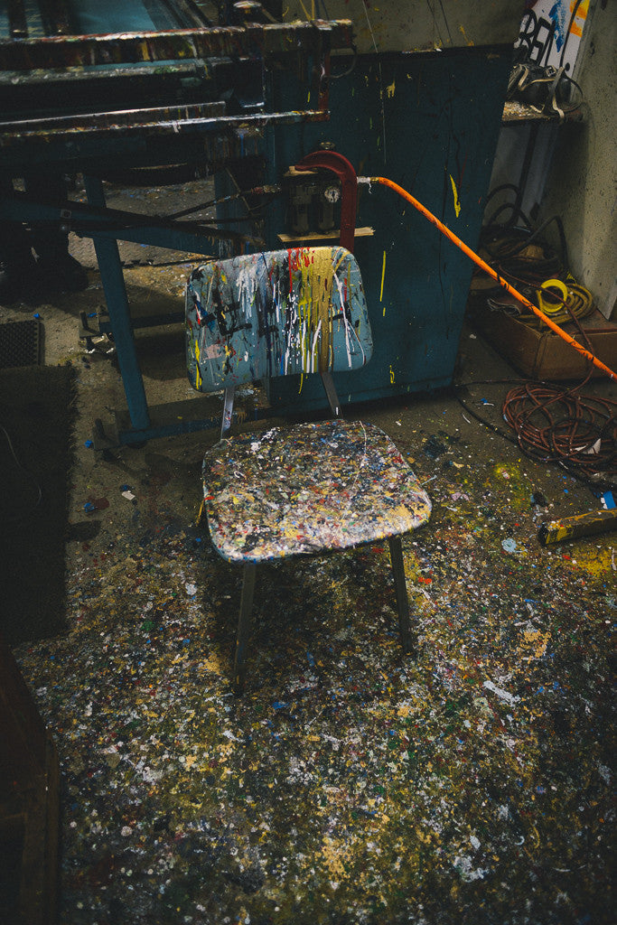 A paint-splattered chair in a cluttered workshop echoes the vibrant chaos of “Chuck’s” by Shaun Roberts with Chuck Sperry. The colorful floor mirrors an archival pigment prints artistry, while tangled cords and industrial machinery complete the lively scene.
