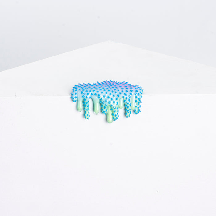 The Mini #14 by Dan Lam is a small abstract sculpture in blue and green, crafted from acrylic and resin with sponge-like spikes. It sits on a white geometric surface, giving a dripping effect against the plain backdrop.