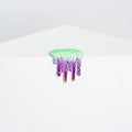 The Mini #23 sculpture by Dan Lam, crafted with hot glue, hangs from a white edge. It showcases a vibrant green spiky top with purple and red drips creating a melting effect against its minimalist background.
