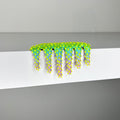 Dan Lam’s "Mini #259," a bright green and yellow spiky caterpillar-like sculpture accented with purple and crafted in mixed media, hangs over the edge of a smooth white surface set against a plain gray background.