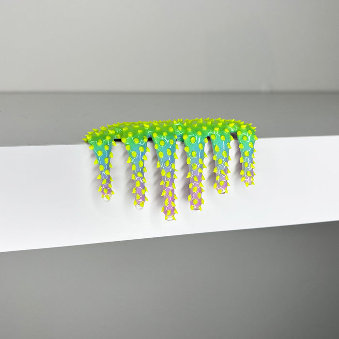 Dan Lam’s "Mini #259," a bright green and yellow spiky caterpillar-like sculpture accented with purple and crafted in mixed media, hangs over the edge of a smooth white surface set against a plain gray background.
