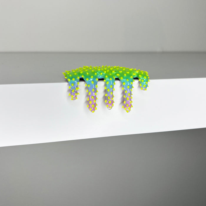 Dan Lam's "Mini #268" is a small, green and blue spiky art object with yellow dots that seems to melt or drip over the edge of a white shelf against a gray background, showcasing the artist’s playful use of mixed media in this 2023 creation.