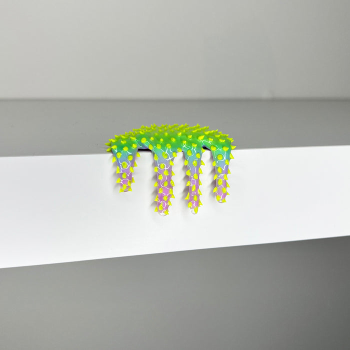 Dan Lams vibrant Mini #278, a 2023-inspired mixed media creation, features a spiky, artificial caterpillar with a gradient from green to purple, hanging over a white edge against a plain gray background.