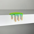 Dan Lam's "Mini #279," a vibrant mixed media art piece featuring colorful, spiky, drippy textures—with yellow and green at the top and blue and purple below—hangs over the edge of a white shelf against a gray background, capturing a striking 2023 aesthetic.
