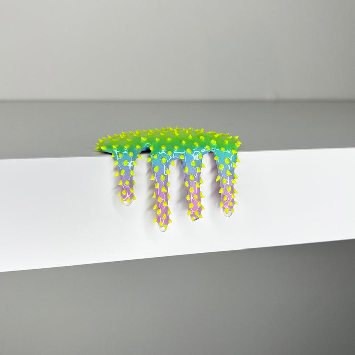 Dan Lam's "Mini #279," a vibrant mixed media art piece featuring colorful, spiky, drippy textures—with yellow and green at the top and blue and purple below—hangs over the edge of a white shelf against a gray background, capturing a striking 2023 aesthetic.