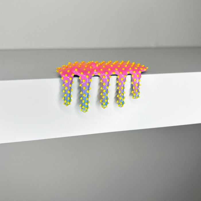 Dan Lams Mini #316, an abstract object with pink, yellow, and purple hues resembling a melting shape, rests vibrantly on a white surface. This striking piece for 2023 showcases bold mixed media brilliance against its plain background.