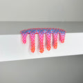 Dan Lam’s "Mini #360," a pink and blue spiked hair clip featuring a drippy mixed media-inspired design, sits on the edge of a white shelf against a plain background.