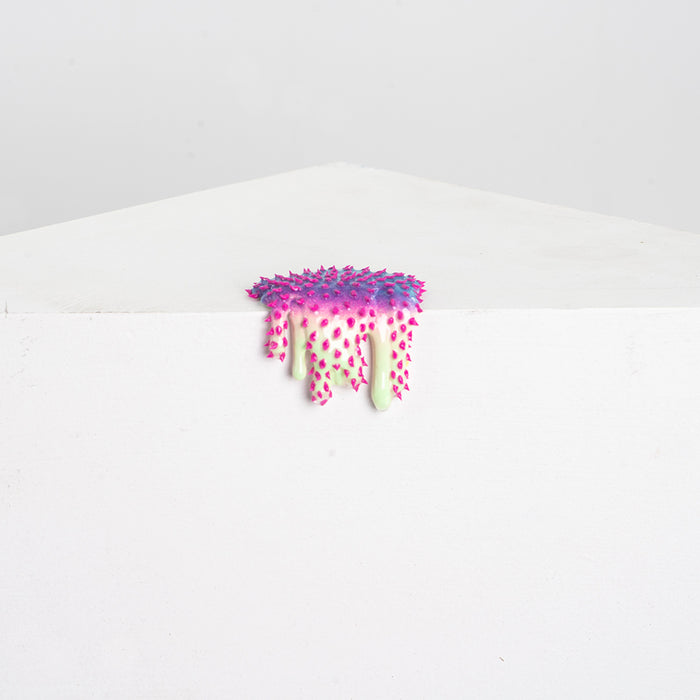The Mini #37 by Dan Lam is a small, colorful resin sculpture resembling a melting scoop of spiky ice cream with vibrant pink and green spikes, perched on a white angular pedestal against a plain gray backdrop.