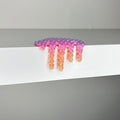 Dan Lam's "Mini #418," a pink and orange star-shaped piece with a dripping effect crafted in mixed media, sits on the edge of a white shelf against a gray background.