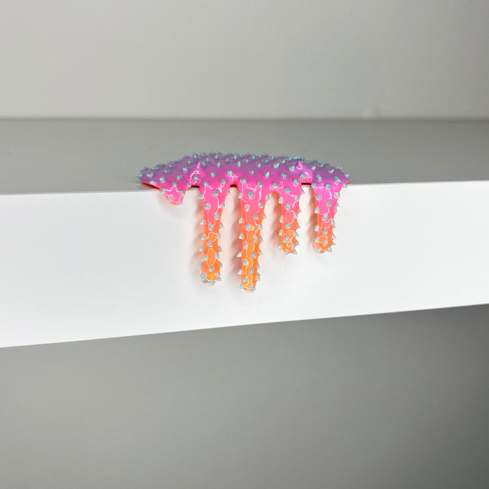 Dan Lam's "Mini #418," a pink and orange star-shaped piece with a dripping effect crafted in mixed media, sits on the edge of a white shelf against a gray background.