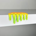 Dan Lam - "Mini #460" by Dan Lam features a yellow and green spiky, slime-like form—crafted with mixed media—dripping over the edge of a white surface against a plain gray background, exemplifying an experimental 2023 aesthetic.