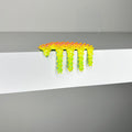 The brightly colored, spiky sculpture Mini #461 by Dan Lam droops over the edge of a white surface like contemporary art, with legs dangling down. The background is out of focus and light gray.
