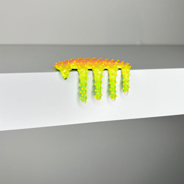 The brightly colored, spiky sculpture Mini #461 by Dan Lam droops over the edge of a white surface like contemporary art, with legs dangling down. The background is out of focus and light gray.