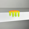 Dan Lams Mini #476 is a small, spiky, multicolored object resembling dripping paint on a white ledge. This striking 2023 mixed media piece transitions from orange to yellow with green tips and appears made of plastic or rubber. The backdrop is a plain gray wall.