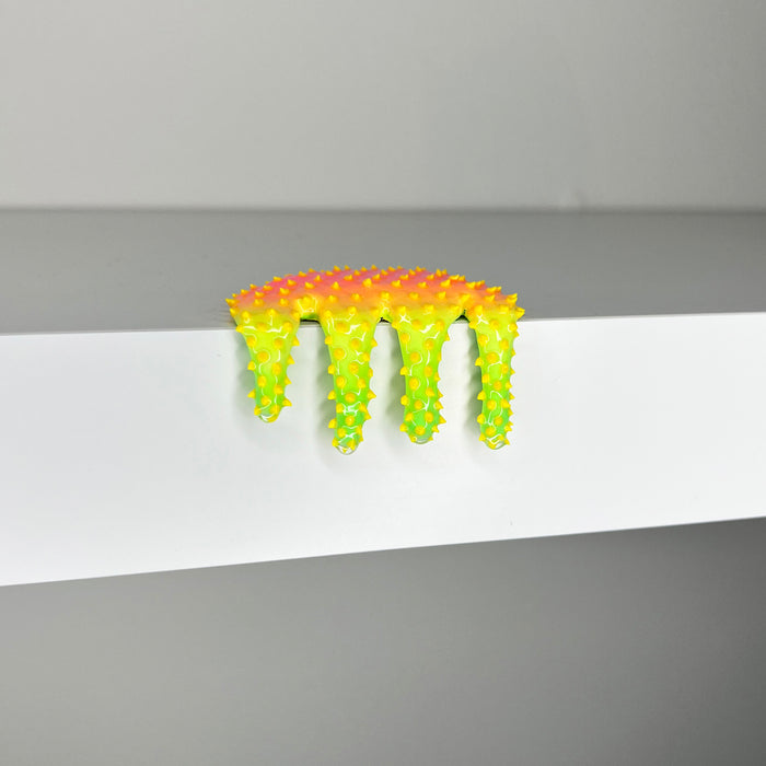 Dan Lams Mini #476 is a small, spiky, multicolored object resembling dripping paint on a white ledge. This striking 2023 mixed media piece transitions from orange to yellow with green tips and appears made of plastic or rubber. The backdrop is a plain gray wall.