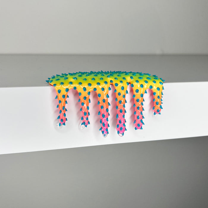 Dan Lam’s "Mini #506" is a captivating mixed media piece from 2023, featuring a vibrant, spiky, slime-like form that drips over the edge of a white surface. Showcasing innovative artistry, the object displays a stunning gradient from yellow to pink, accented by blue spikes tipped with small white spheres.