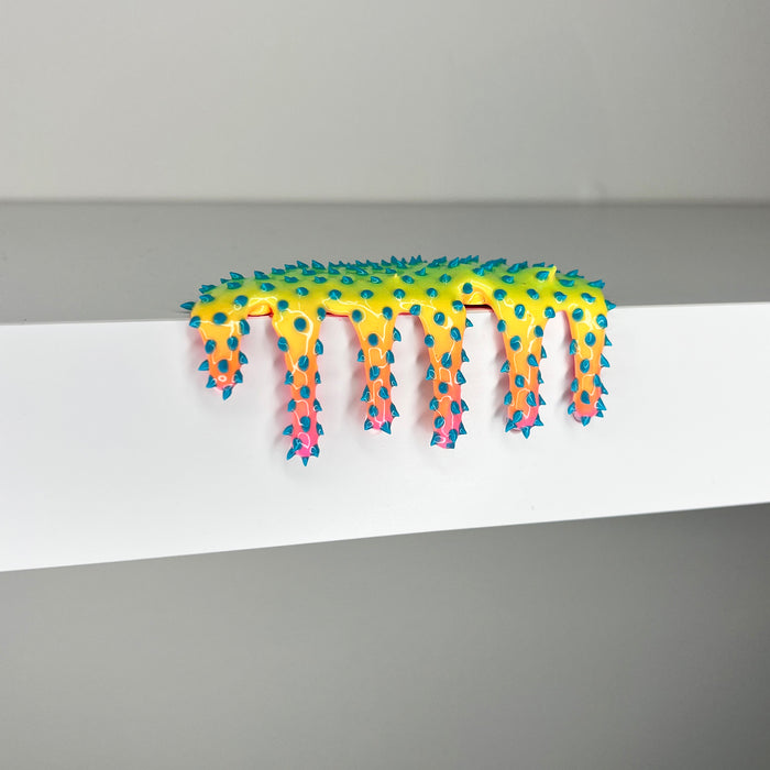 Dan Lams Mini #510 features a spiky, neon yellow and orange design with blue tips dripping over a white shelf against a plain backdrop. Reminiscent of 2023 mixed media art, its colorful and abstract style blends liquid and spiked textures.