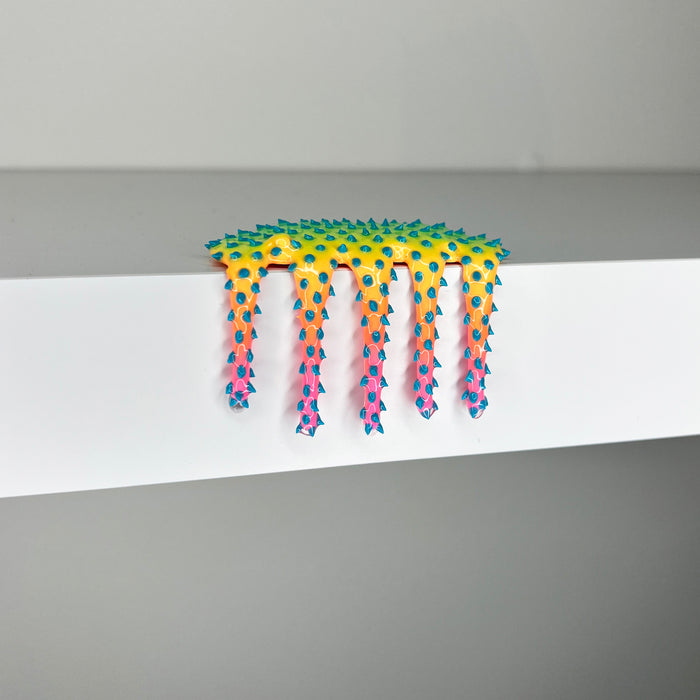 Mini #520" by Dan Lam is a colorful mixed media sculpture featuring vibrant yellow and green hues, with pink and blue-tipped spikes resembling dripping slime. Hanging over the edge of a white surface against a gray background, this striking 2 x 3 x 1 3/4 inch artwork was created in 2023.
