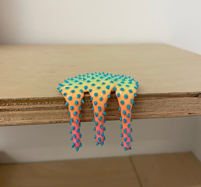 Dan Lam's "Mini #539" is a colorful mixed media piece from 2023, featuring spiky blue protrusions and a yellow-to-pink gradient that drips over the edge of a wooden shelf.