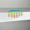 Dan Lams 2023 mixed media piece, Mini #558, displays a colorful, wavy-edged form resembling an abstract comb that drips over a white surface. The gradient transitions from blue to green to orange and creates a soft, jagged texture.
