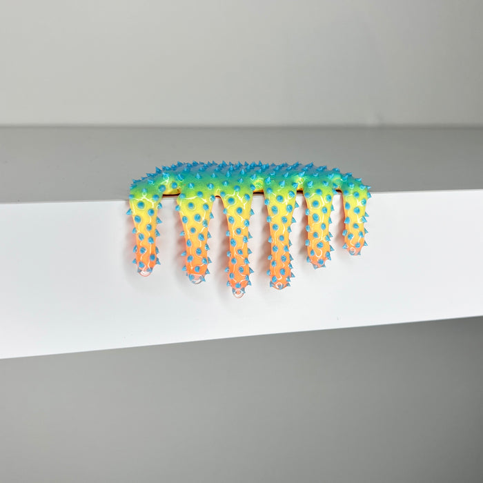 Dan Lams 2023 mixed media piece, Mini #558, displays a colorful, wavy-edged form resembling an abstract comb that drips over a white surface. The gradient transitions from blue to green to orange and creates a soft, jagged texture.