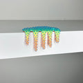 Dan Lams Mini #569 from 2023 showcases colorful, spiky silicone polymer dripping over a white shelf. This mixed-media art features a gradient of green, yellow, orange, and pink with spike-like textures against a neutral background.