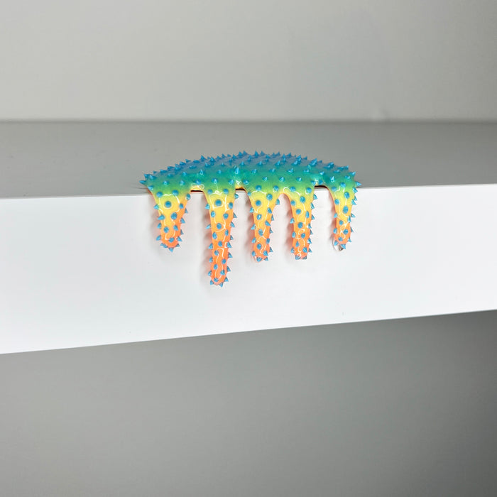 Dan Lam’s "Mini #570" is a colorful, spiky, blob-like mixed media sculpture featuring dramatic drips that spill over the edge of a white surface against a plain gray background.