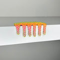 The Mini #58 by Dan Lam, a vibrant cactus-shaped comb with green spikes, sits on a white shelf. This 2023 design masterpiece blends pink to orange gradients in mixed media, making it both functional and visually stunning.