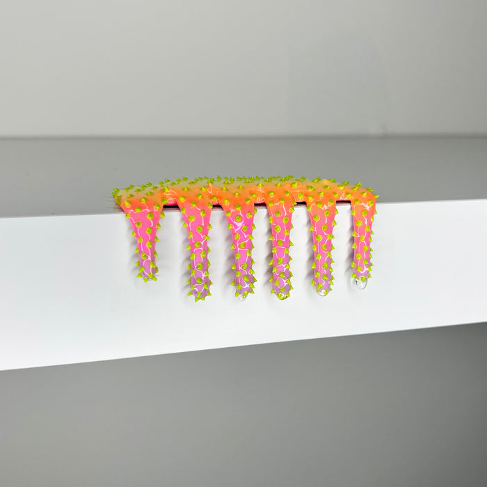 The Mini #58 by Dan Lam, a vibrant cactus-shaped comb with green spikes, sits on a white shelf. This 2023 design masterpiece blends pink to orange gradients in mixed media, making it both functional and visually stunning.