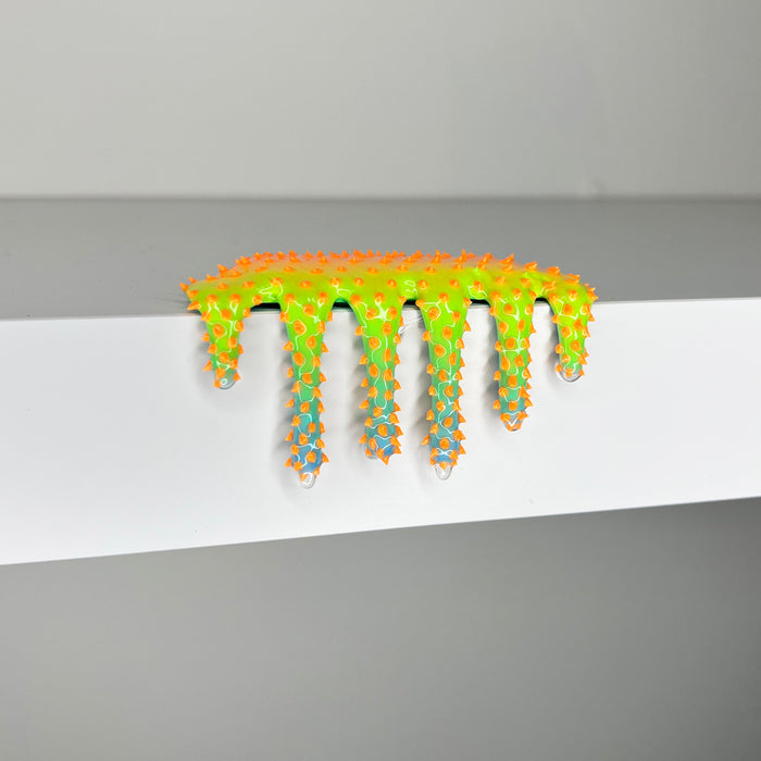 Dan Lam's "Mini #609" is a 2023 mixed media piece featuring neon green and orange goo-like forms with blue accents and spiked protrusions, dripping over the edge of a white shelf. This striking artwork from Dan Lam blends bold color and texture for an eye-catching effect.
