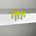 Dan Lams Mini #620, a vibrant mixed media sculpture, resembles melting slime and sits on a white shelf. It has a bright green surface with orange and blue star-like protrusions, offering a textured, playful look that feels refreshingly 2023.