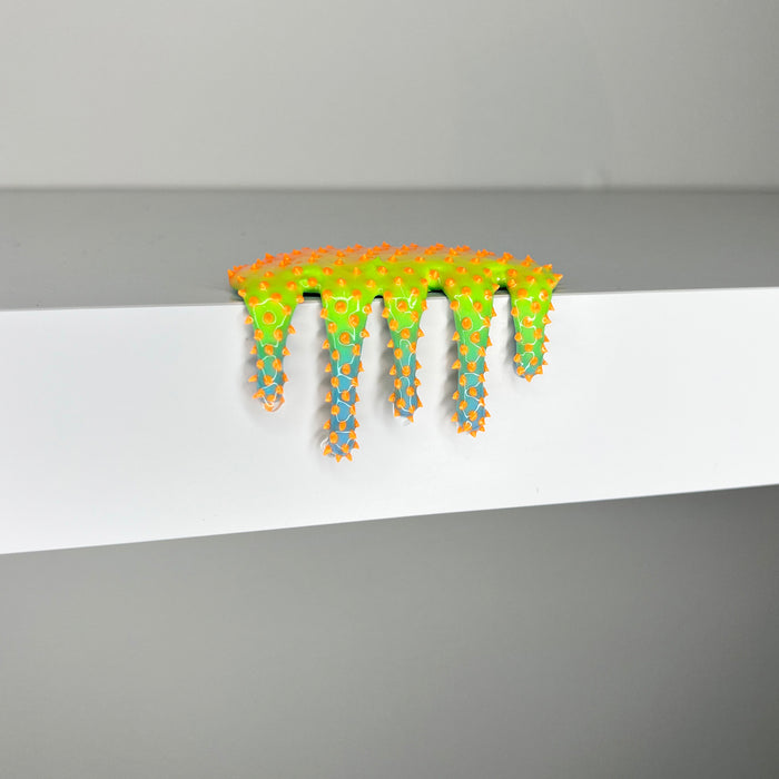 Dan Lams Mini #620, a vibrant mixed media sculpture, resembles melting slime and sits on a white shelf. It has a bright green surface with orange and blue star-like protrusions, offering a textured, playful look that feels refreshingly 2023.