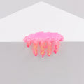 Dan Lams Mini #7.05, a small pink jelly-like shape with dripping fringes, rests on the edge of a white geometric surface. Resembling a cloud or jellyfish and crafted from transparent resin, it stands out against the plain gray background.