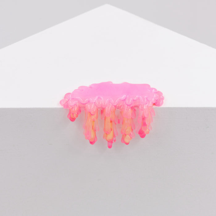 Dan Lams Mini #7.05, a small pink jelly-like shape with dripping fringes, rests on the edge of a white geometric surface. Resembling a cloud or jellyfish and crafted from transparent resin, it stands out against the plain gray background.